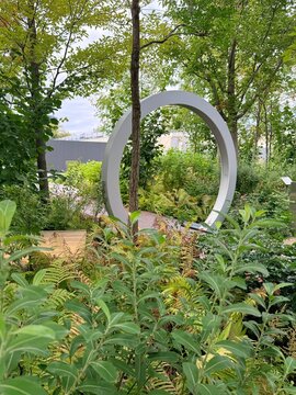 Large grey metal ring sculpture in a lush green garden with ferns, trees, and wooden pathways, representing modern landscape architecture.