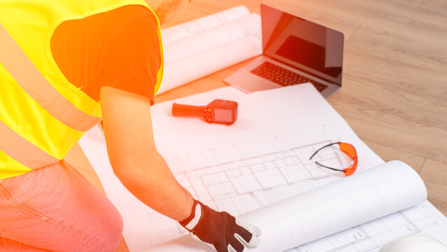 Worker wears yellow safety vest. Hand holds pen over blueprints. Laptop and tape measure sit nearby. Worker focuses on construction plans. Professional setting for design work
