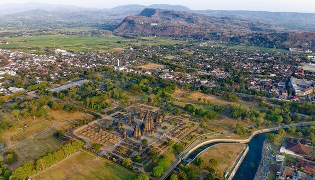Aerial view of Prambanan Temple surrounded by ancient stone structures, green grounds, and nearby urban areas with mountains in Kecamatan Prambanan, Daerah Istimewa Yogya, Indonesia.