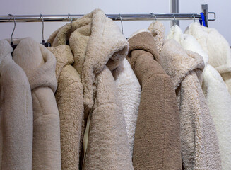 Collection of soft shearling and teddy fleece coats in neutral beige and white tones hanging on metal hangers and a clothing rack in an indoor wardrobe setting © ravital