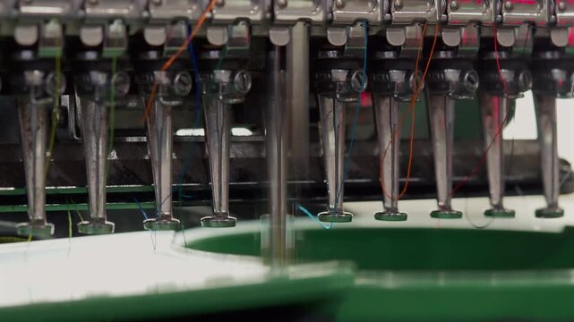 A detailed industrial shot of a multi-head embroidery machine. Multiple needles with colorful threads are poised above a blue fabric taut in an embroidery hoop.