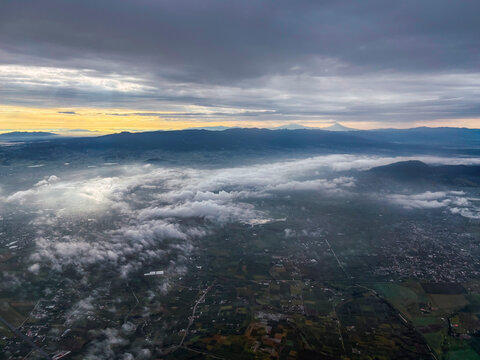 Aerial photograph of a landscape in Ixtlahuaca, Mexico, at sunrise, featuring clouds and a sky with very warm tones; Santa Cruz Hill is visible, with Popo and Izta in the background.
