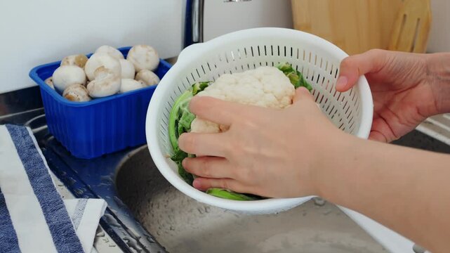 Person washes raw cauliflower inside plastic strainer over modern kitchen sink, carefully holding vegetable beneath flowing tap water stream during thorough fresh ingredient rinsing routine.