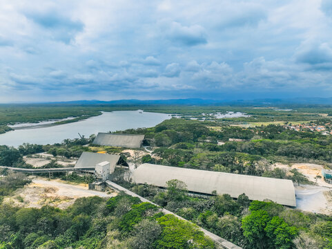 Aerial view of an industrial facility with large arched warehouses and conveyor belts situated near a river and lush forest in Cilacap, Central Java, Indonesia.