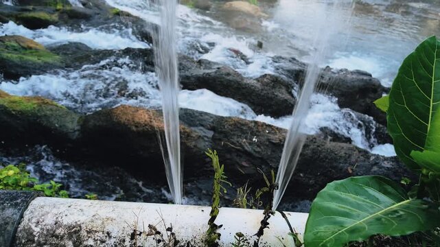 Water spraying from a leaking white pipe in front of a rushing rocky stream surrounded by green plants