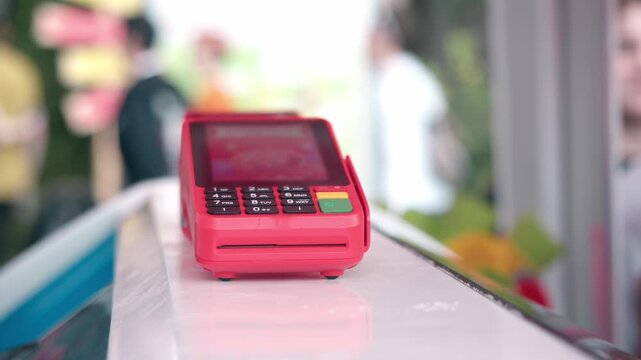 Close up shows red wireless payment terminal sitting on white counter of outdoor park food kiosk. modern pos machine waits for credit card transactions.