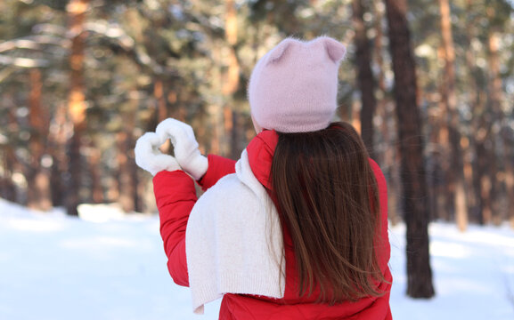 Girl in red jacket shows heart with hands in snowy winter forest, winter walk in the forest. Back view, young woman in pink hat makes heart gesture with gloves on blurred trees background. Nature
