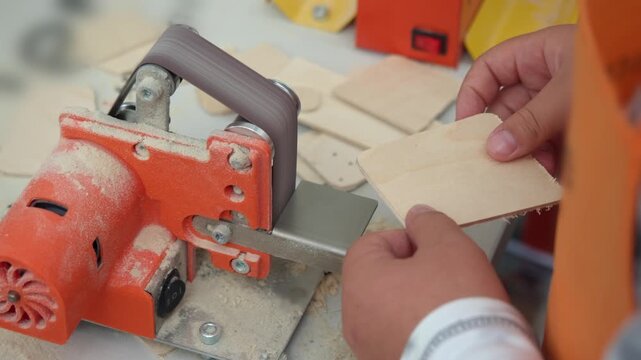 Child guides light wood piece against small electric belt sander in workshop. Bright orange power tool creates sawdust while shaping craft project.