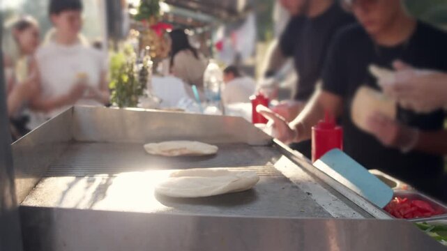 Gloved chef hand removes baked flatbreads from hot metal griddle. Busy worker prepares fresh warm street food for hungry festival crowds waiting outside in bright summer sunlight.