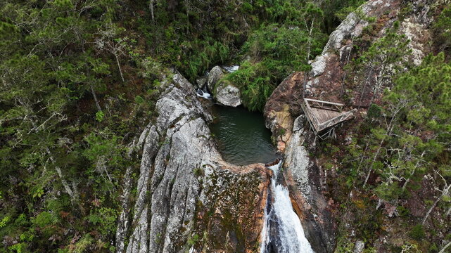 Aerial view of Salto de Aguas Blancas waterfall cascading over rocky cliffs surrounded by lush pine forest and a wooden observation deck in Constanza, La Vega Province, Dominican Republic.