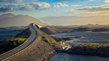 Lyngholmen, Norway - 23.06.2024: Curved asphalt road leading to a concrete bridge over blue sea with misty mountains under golden hour light © Andrei