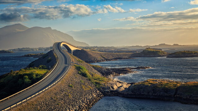 Lyngholmen, Norway - 23.06.2024: Curved asphalt road leading to a concrete bridge over blue sea with misty mountains under golden hour light