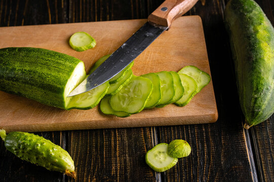 Kitchen knife cuts cucumber into rounds on wooden board. Fresh cucumber pieces and whole vegetables frame cutting area, highlighting everyday food preparation against dark wooden surface.