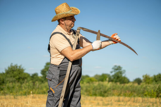 A worker in a straw hat stands in a field holding a scythe. He adjusts the tool while looking at the surrounding area. The sun shines brightly in the sky