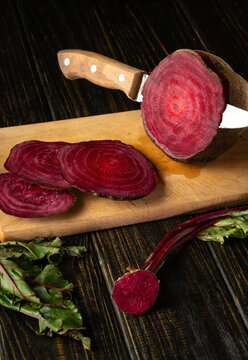 Beetroot is cut into round slices on wooden cutting board, with knife handle visible and leafy stems around it. Rich red flesh and rustic styling give this food prep image warm, homely mood.