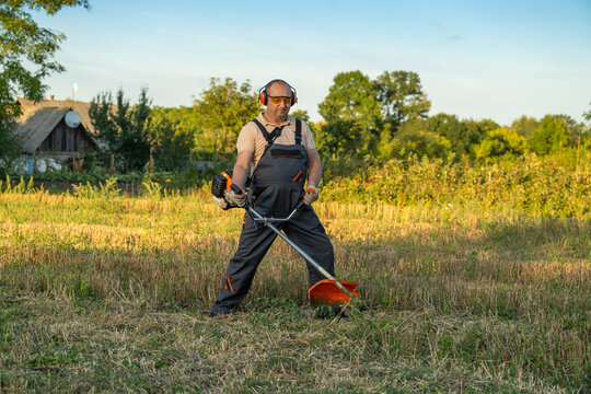 Man using brush cutter in field, wearing ear protection and work overalls while trimming dry grass. He stands focused in rural summer light near house and trees, doing outdoor grounds maintenance.