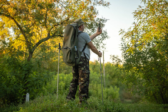 Backpacker stands still on woodland path, raising smartphone for self portrait. Man with hiking poles and camouflage pants appears content in sunlit autumn greenery beside dense trees.