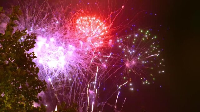 Bright colors fill the sky as fireworks burst above the city. The event takes place during the night hours. People gather in parks and streets to watch the show and enjoy the moment together.