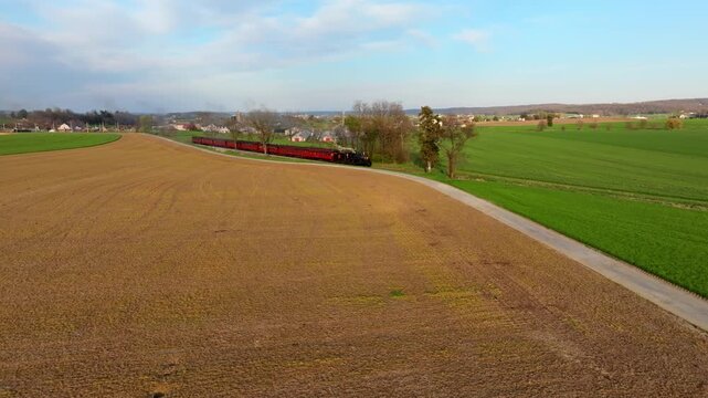A train moves steadily along its tracks through open fields and rolling hills. The landscape features patches of green grass and harvested brown fields under a blue sky.