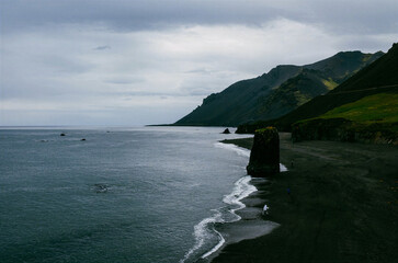 Dark green mountain coastline meets calm water beneath an overcast sky, creating a remote scenic landscape ideal for travel, nature and dramatic coastal themes. © UQink
