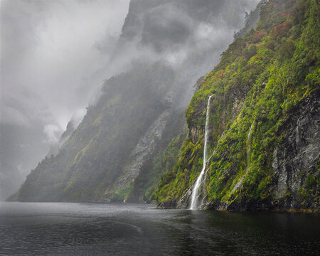 Steep green mountain cliffs disappear into mist as a narrow waterfall drops through the valley, creating a dramatic wilderness scene ideal for travel, hiking and nature designs.