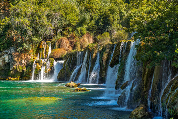 Stunning landscape of the Kravice Waterfall near Mostar, Bosnia and Herzegovina. Multi-tiered cascades flowing into a turquoise emerald lake surrounded by lush green trees and mossy cliffs © Yuliko