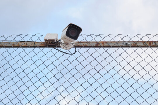 Security camera mounted on metal chain link fence under cloudy sky view