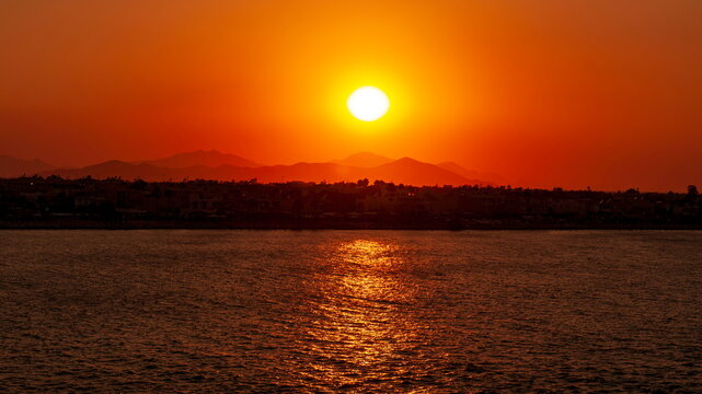 Kos, Greece - 20.07.2014: Golden sunset over Aegean Sea with silhouettes of coastal buildings and distant mountains under a warm orange sky