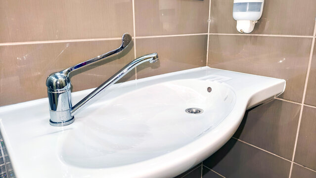 White bathroom sink with long lever faucet against tiled wall. Wall mounted soap dispenser for handwashing. Bathroom fixture, hygiene and sanitation. Washroom interior and plumbing