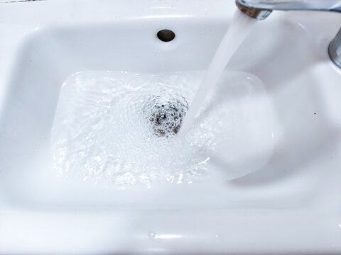 Faucet running into white bathroom sink as water swirls down the drain. Household plumbing and sanitation. Fresh water use and hygiene concepts. Bathroom fixture for domestic cleaning