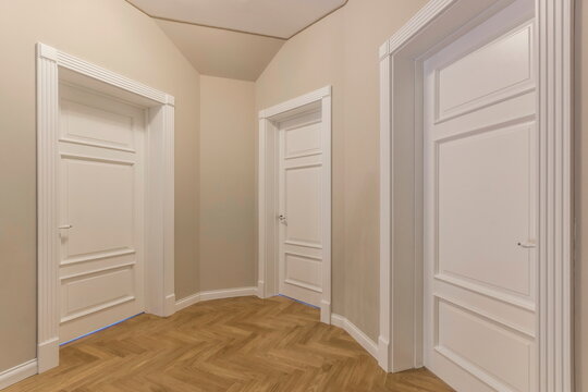 A hallway features three white doors aligned along the walls. The flooring is made of light wood, creating a simple residential space. The walls are