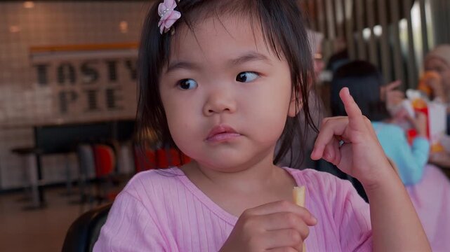 Young Asian toddler girl eating at a fast-food restaurant, hand near her mouth.
