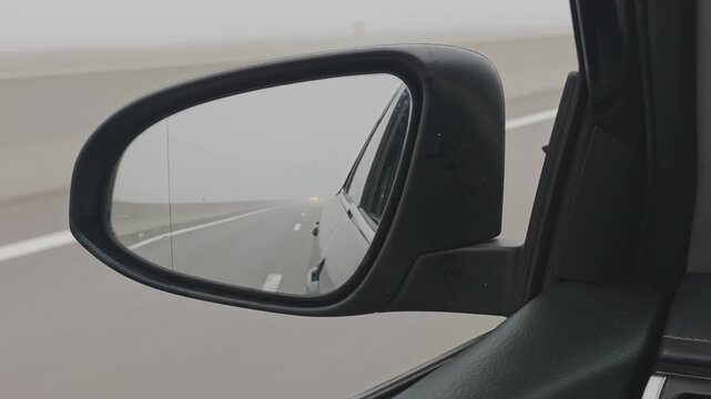 Driver's point of view from a car traveling on a highway during a foggy day. The side mirror reflects the road with low visibility