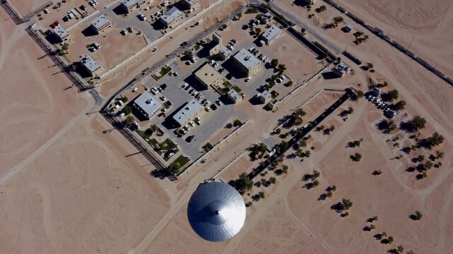 Aerial desert dome facility with metallic radome and surrounding research compound, sunlit sandy terrain, grid roads, scattered buildings, palm clusters, perimeter fence, service vehicles, drone