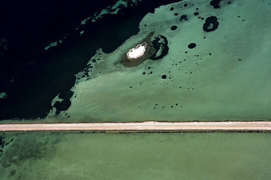 Aerial view of the causeway crossing the shallow turquoise and dark blue waters near Useless Loop, showcasing salt mining patterns and a small island in Kalbarri, Western Australia, Australia.