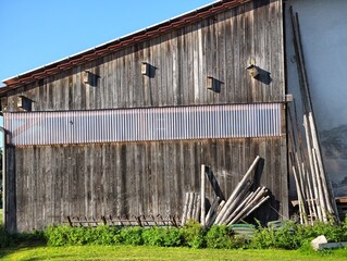 Weathered wooden barn wall with stacked poles beside farmyard under blue sky © Waldemar