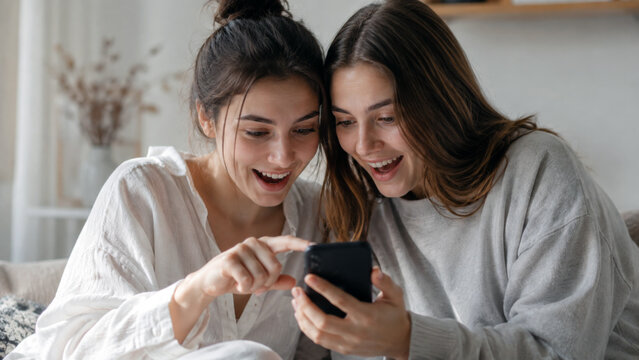 Two young women smiling and sharing content on a smartphone together while relaxing at home