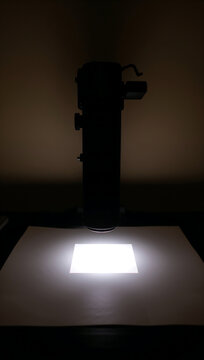 A dark silhouette of a vintage photographic enlarger casting a bright square of light onto a white paper surface in a dark room for processing