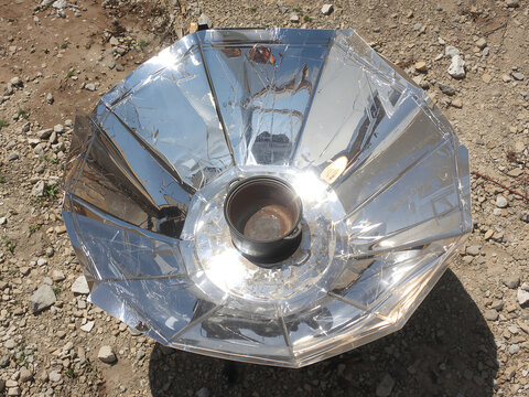 A solar cooker with a reflective metallic parabolic dish and a central black cooking pot, positioned on dry rocky ground under bright sunlight rays
