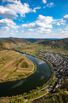 Aerial view of a river loop winding through terraced vineyards and a small town under a bright blue sky in Kelberg, Rhineland-Palatinate, Germany.