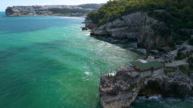 Trabucco di Monte Pucci aerial wide shot flying along rocky coast towards Peschici town, Gargano Puglia Italy over turquoise Adriatic Sea, cinematic drone flight along jagged coastline