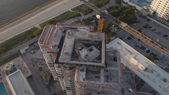 Aerial view of a high-rise building under construction with a yellow crane, adjacent parking lot, and coastal scenery in Miami Beach, Florida, United States.