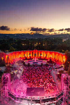 Aerial view of Park Nazzjonali Ta' Qali during an outdoor concert with a large crowd, vibrant red lighting, a central fountain, and Mdina skyline in Attard, Northern Region, Malta.