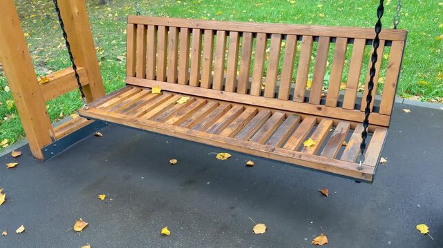 Wooden porch swing on chains in a public park with scattered autumn leaves. Outdoor furniture background, cozy backyard garden design, empty recreation area, peaceful fall season.