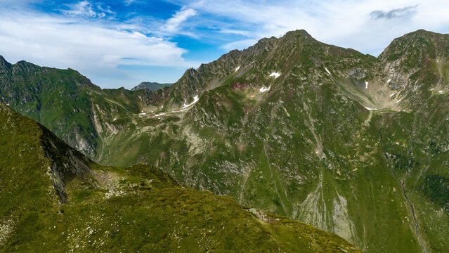 Aerial view of the Transfagarasan mountain pass, showcasing rugged green peaks, rocky slopes, and patches of snow under a blue sky in Curtea de Arges, Judetul Arges, Romania.