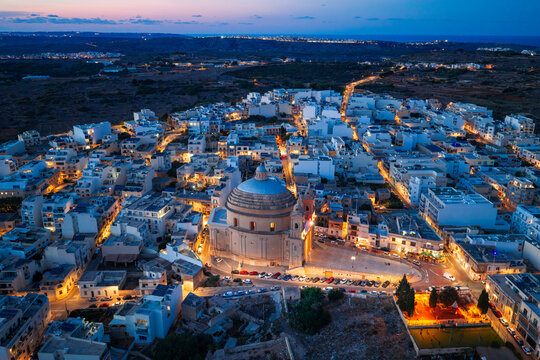 Aerial view of the Mgarr Parish Church illuminated at night, surrounded by the dense urban fabric of the town, with warm street lights glowing in Mgarr, Northern Region, Malta.