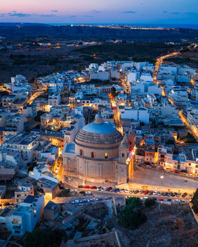 Aerial view of the Parish Church of the Assumption of the Blessed Virgin Mary into Heaven illuminated at twilight, surrounded by the dense urban landscape of Mgarr, Northern Region, Malta.