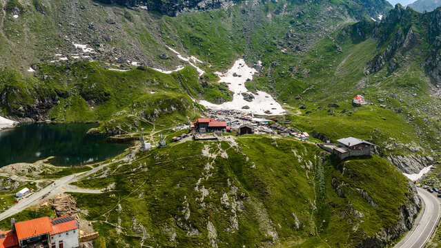Aerial view of Transfagarasan mountain pass featuring Balea Lake, mountain chalets, winding roads, and snow patches on the green slopes in Curtea de Arges, Judetul Arges, Romania.