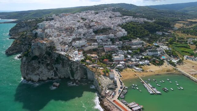 Gargano Puglia Italy Aerial flyby reveal of Peschici white town marina and beach sideways drone flight along rocky coastline with umbrellas and waves. Italian summer cinematic b-roll 4k