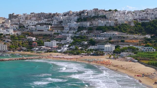 Panoramic view of Peschici white town on rocky cliff coast Gargano Puglia Italy cinematic drone 70 mm over Adriatic Sea showing town beach and coastline